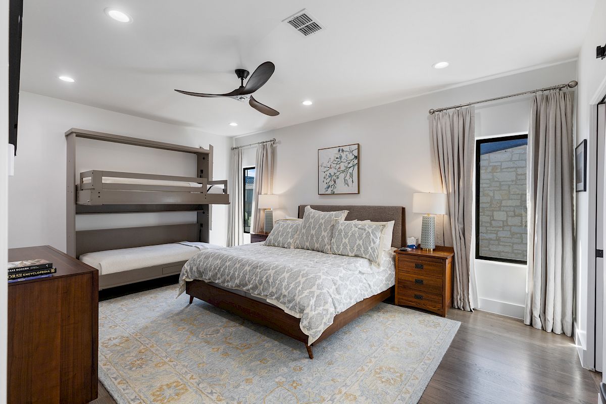 A modern bedroom with a large bed, gray quilt, ceiling fan, nightstands, lamps, and a Murphy bed. Curtains cover windows, adding elegance.