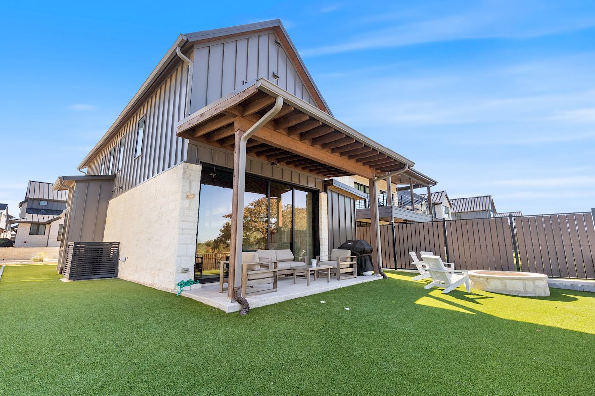 A modern house with a patio, outdoor furniture, and artificial grass. There's a small fire pit and clear skies in the background.