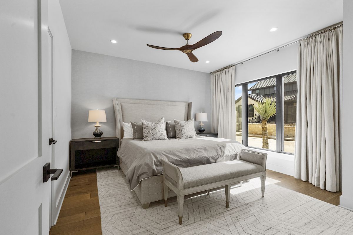 A modern bedroom with a large bed, bench, side tables with lamps, ceiling fan, and a window with drapes.