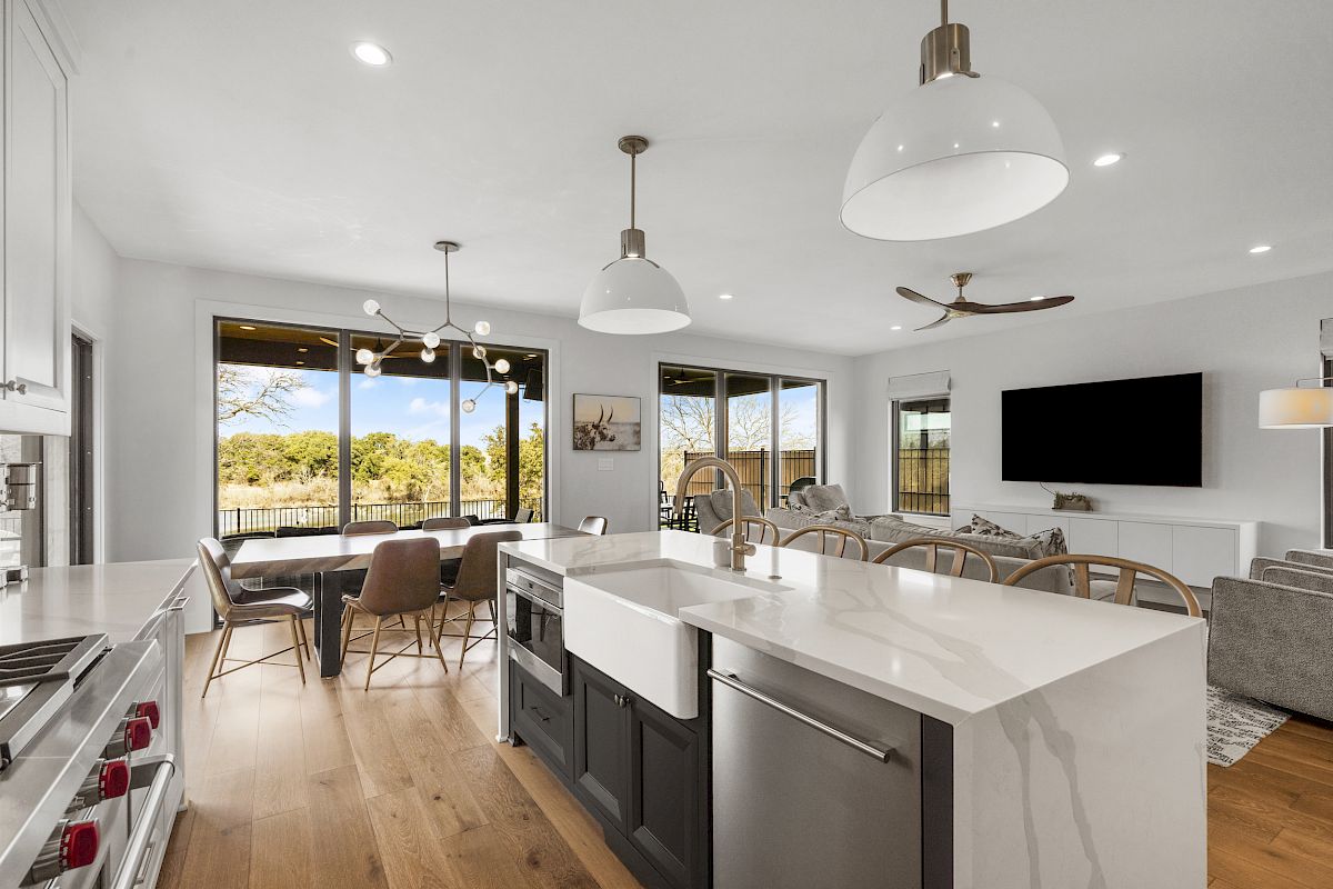 A modern kitchen and living area with an island, pendant lights, and a large TV, featuring a sleek design and outdoor view.