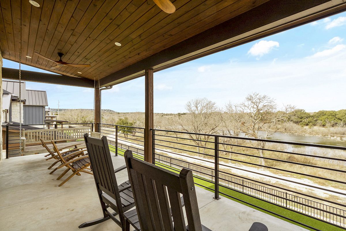 A covered outdoor deck with wooden rocking chairs overlooks a scenic view of trees, a river, and the sky in the background.