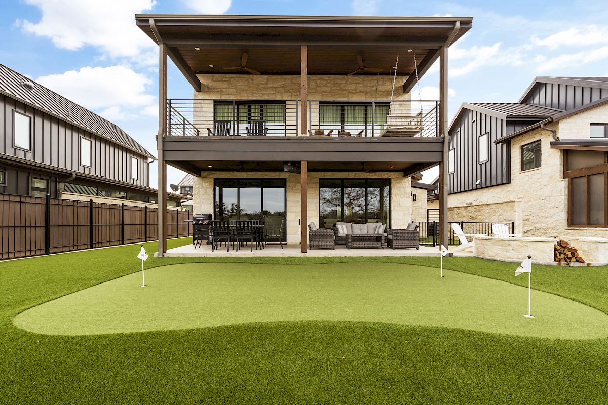 A modern house with a large balcony and a backyard putting green surrounded by well-maintained grass and patio seating.