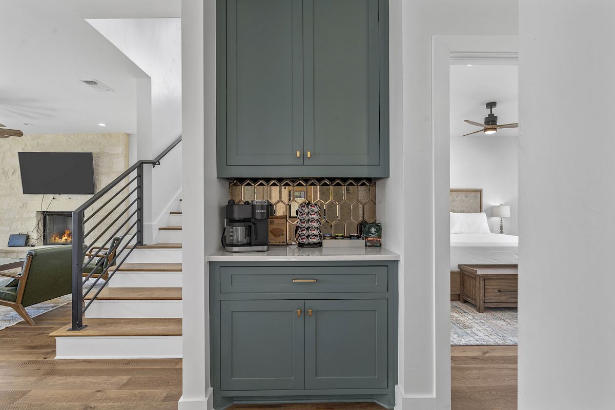 The image shows a small coffee station in a home, with teal cabinets, a coffee maker, and a view into a living room with a TV and stairs.