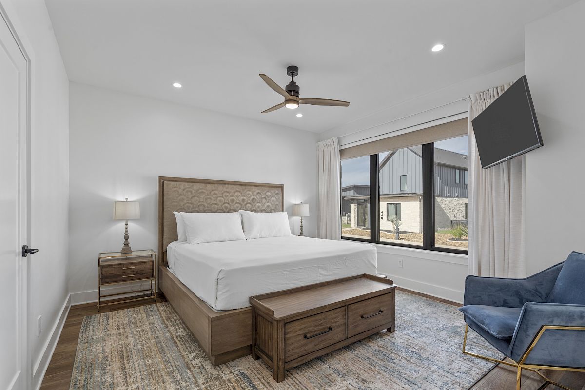 A modern bedroom with a bed, nightstands, a ceiling fan, a wall-mounted TV, large window, and a blue chair on a patterned rug.