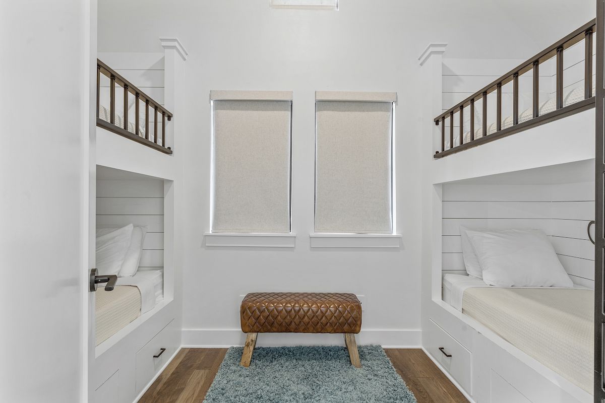The image shows a room with two sets of bunk beds, a small bench, and a blue rug, featuring white walls and wooden floors.