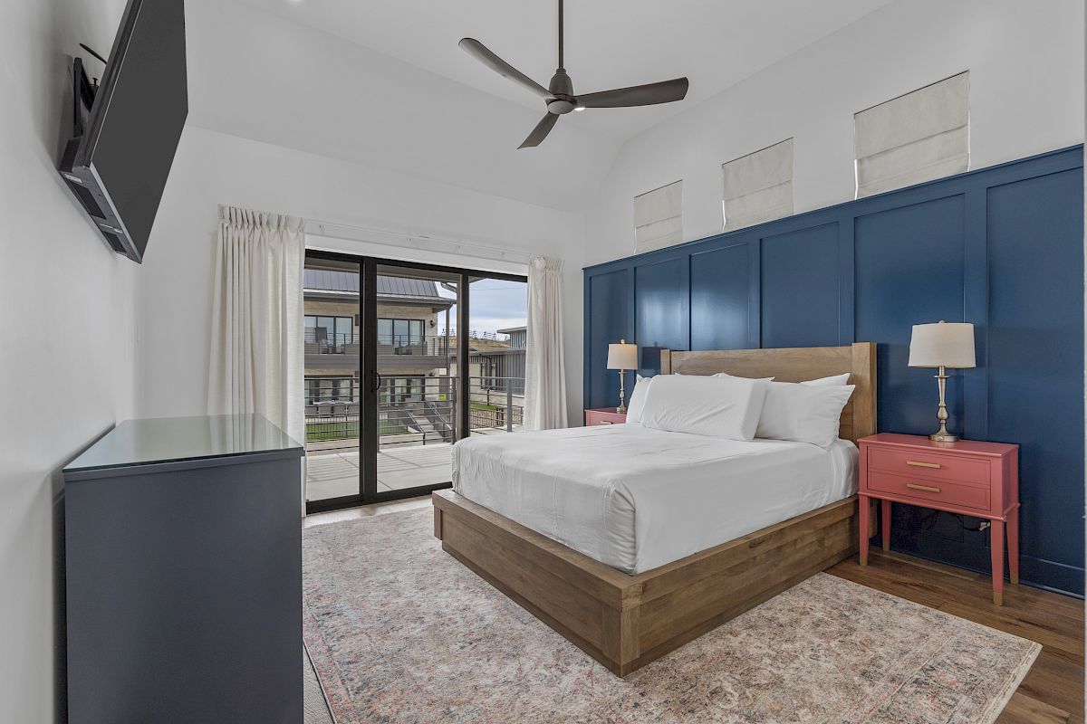 A bedroom with a bed, blue accent wall, pink side tables, lamps, TV, ceiling fan, rug, and sliding doors leading to a balcony outside.