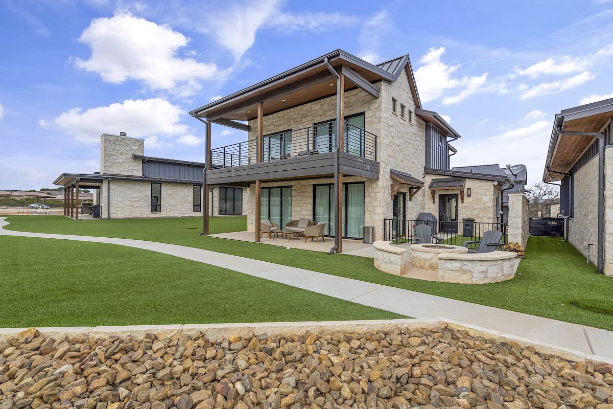 A modern house with a two-story design, large windows, and stone walls. There's a small patio area, green lawn, and a stone fire pit.