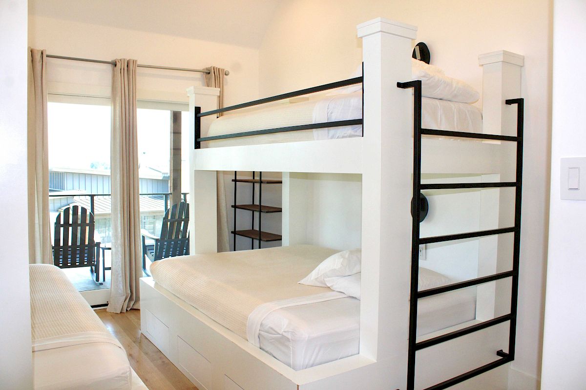 A room with white bunk beds, a ladder, shelves, and a sliding glass door leading to a balcony with chairs outside.