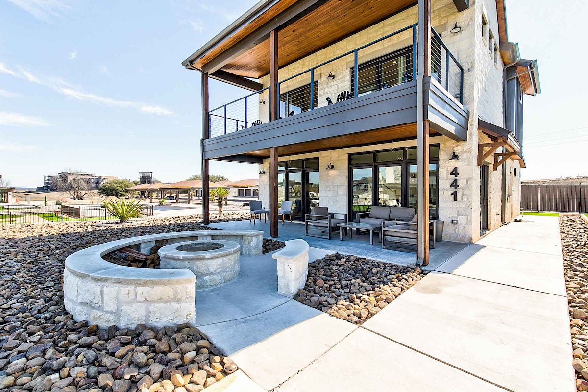 A modern two-story house with a stone exterior, balcony, outdoor seating area, and fire pit surrounded by gravel and landscaped paths.