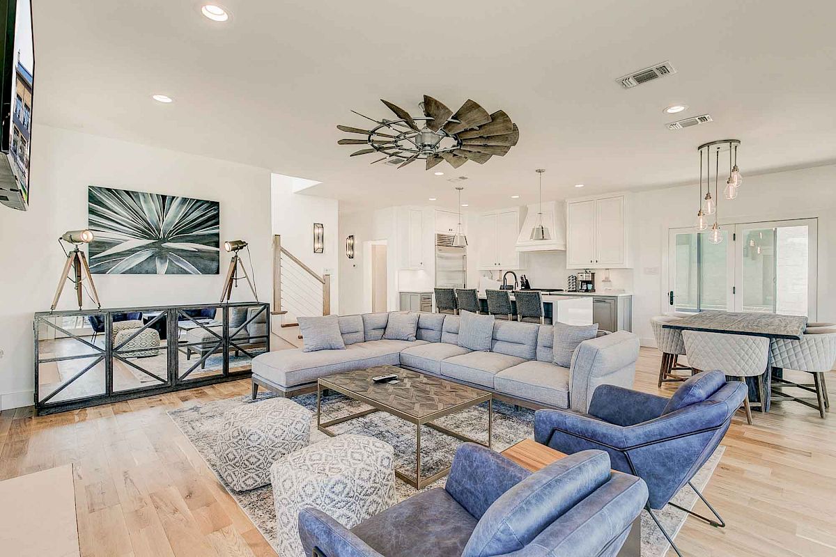 The image shows a modern living room with a sectional sofa, blue armchairs, stylish decor, and an open kitchen area.