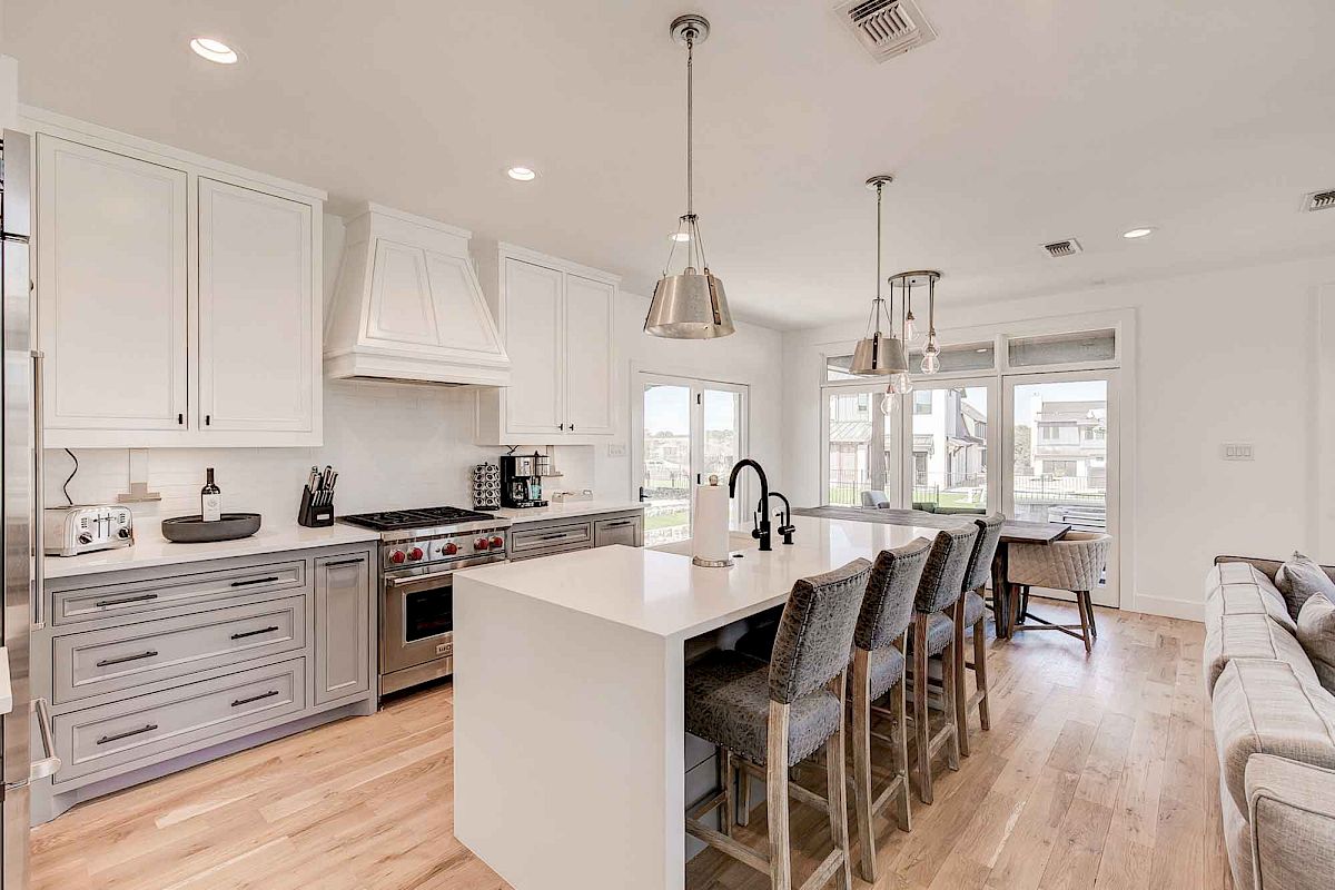 A modern kitchen with white cabinets, island, bar stools, and pendant lights. Large windows offer natural light and outdoor views.