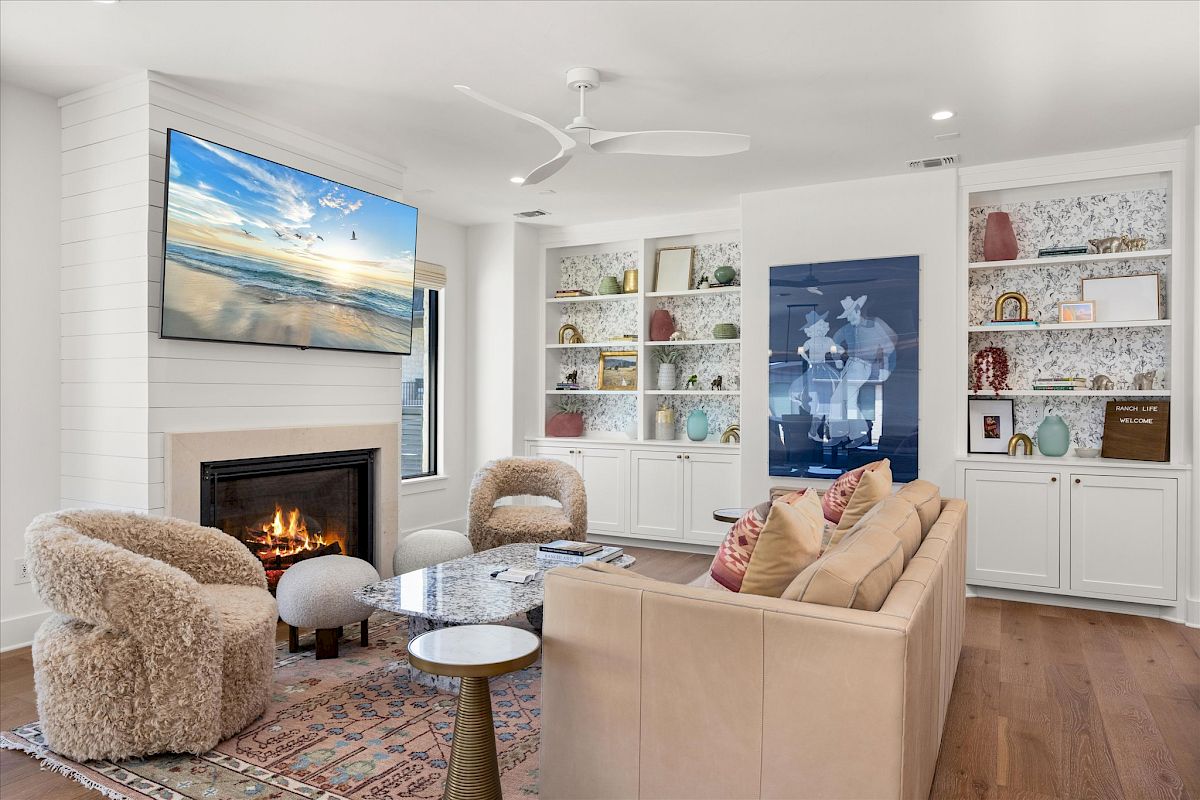 A cozy living room with a beige sofa, fluffy chairs, fireplace, wall art, and a TV above. Bookshelves display various decor items.
