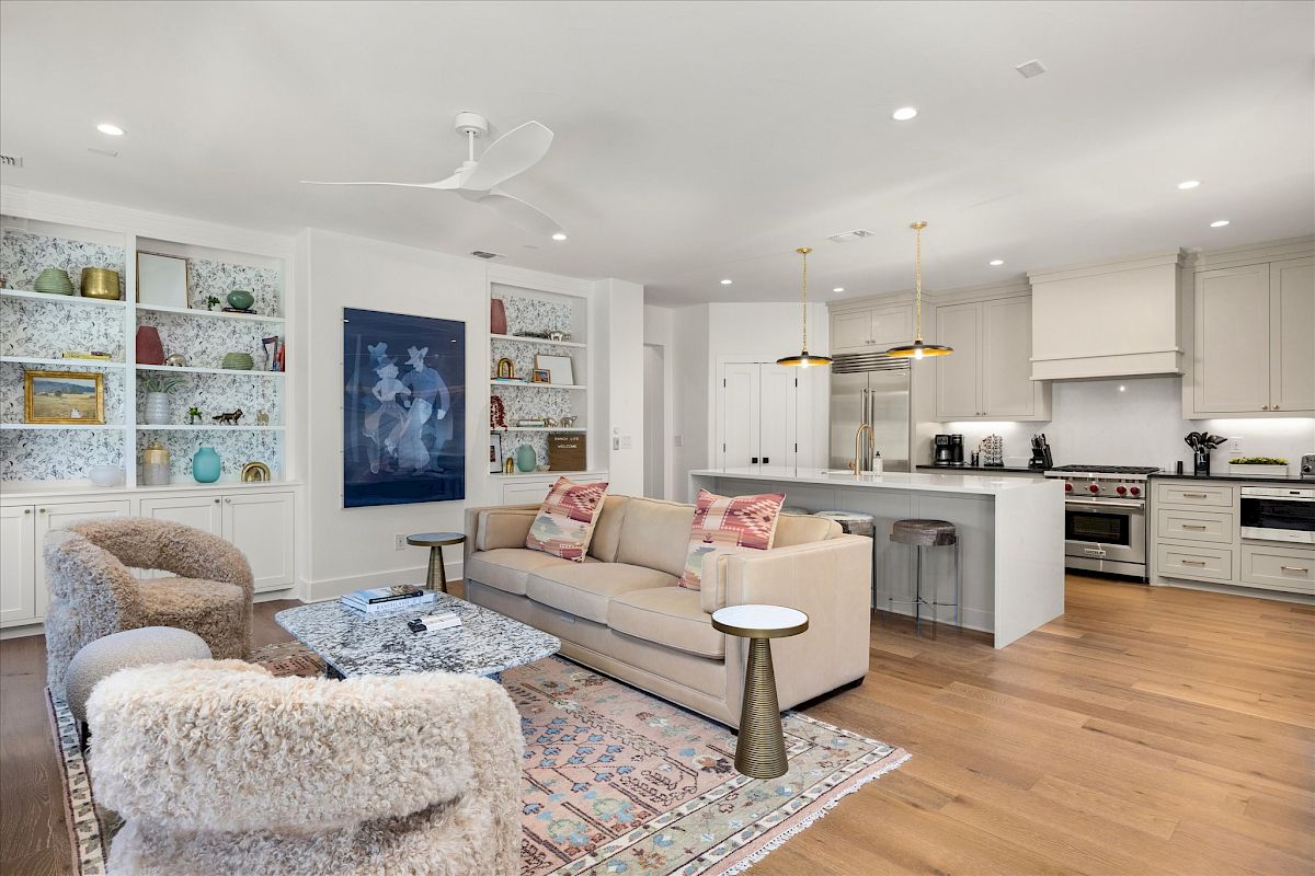 A modern living room with a sofa, armchairs, coffee table, wall art, open shelves, and a kitchen with appliances in the background.