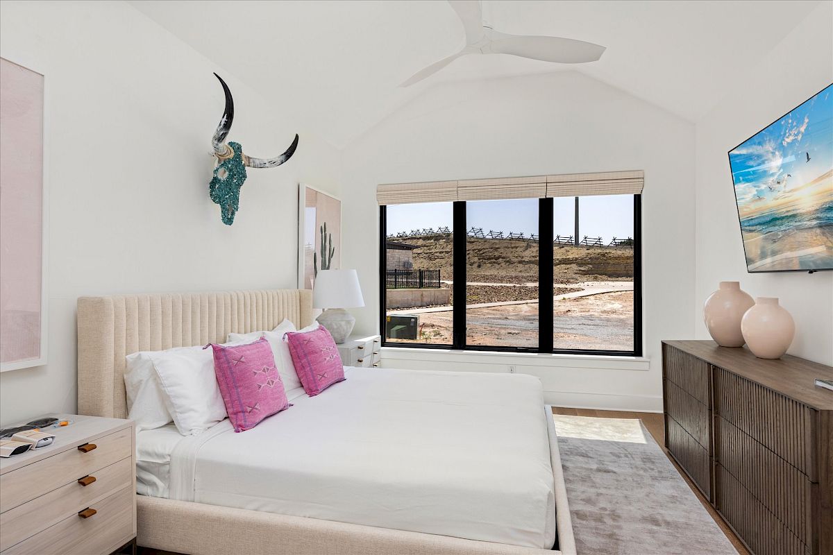 A modern bedroom features a bed with pink pillows, a wall art piece, large window, wooden furniture, and a wall-mounted TV.