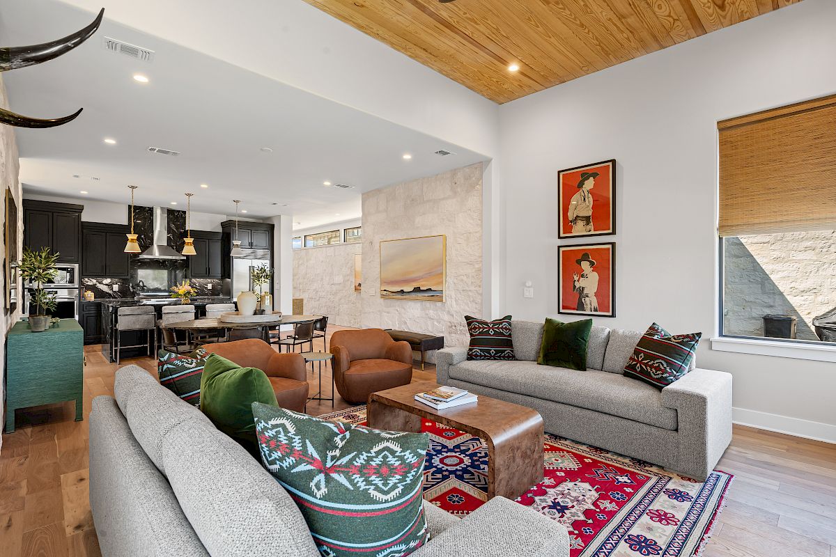 A modern living room with gray sofas, colorful cushions, a wooden ceiling, art on the walls, and an adjoining kitchen and dining area.