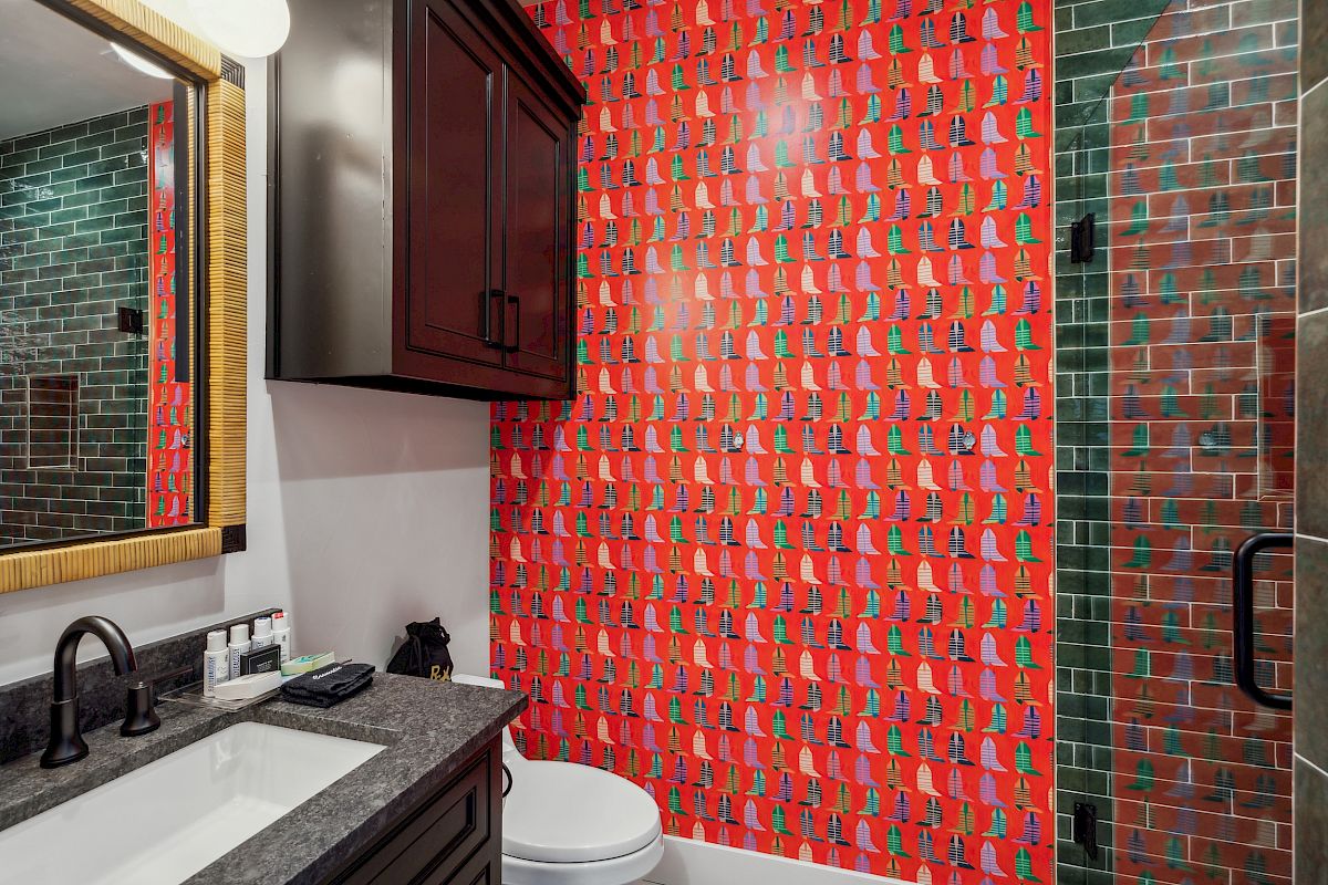 A bathroom with a red patterned wall, dark wood cabinet, green-tiled shower, and a sink with black fixtures and toiletries.