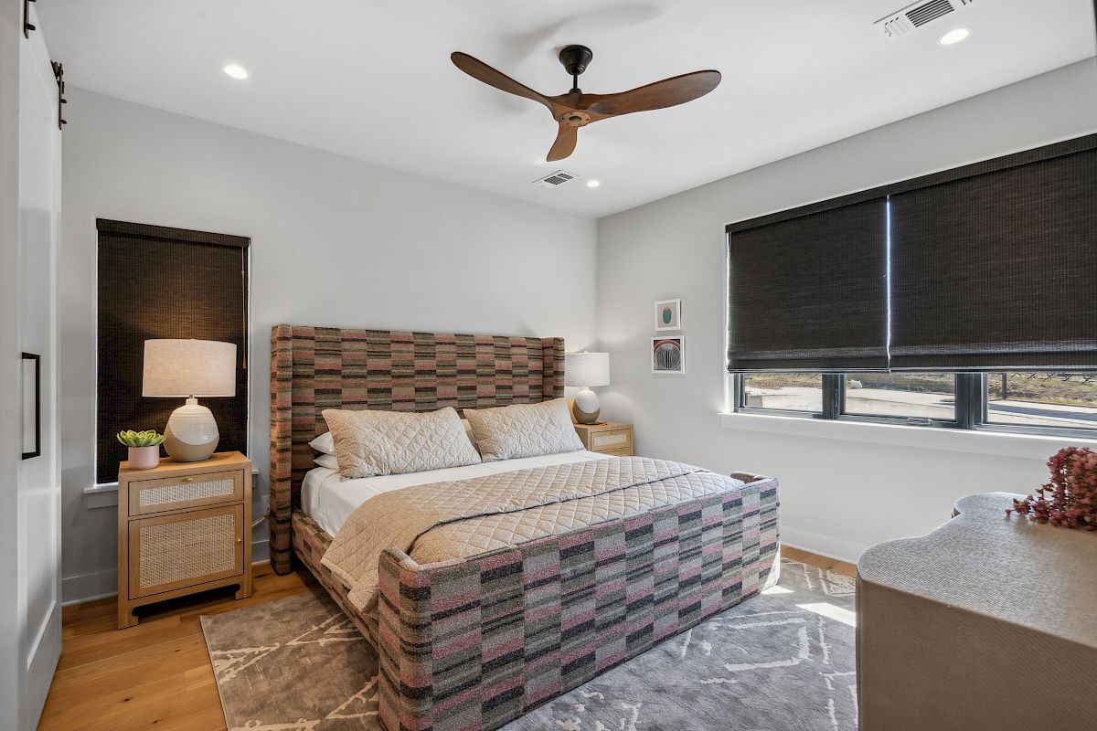 A modern bedroom features a large bed, two side tables with lamps, a patterned rug, closed blinds, and a ceiling fan.