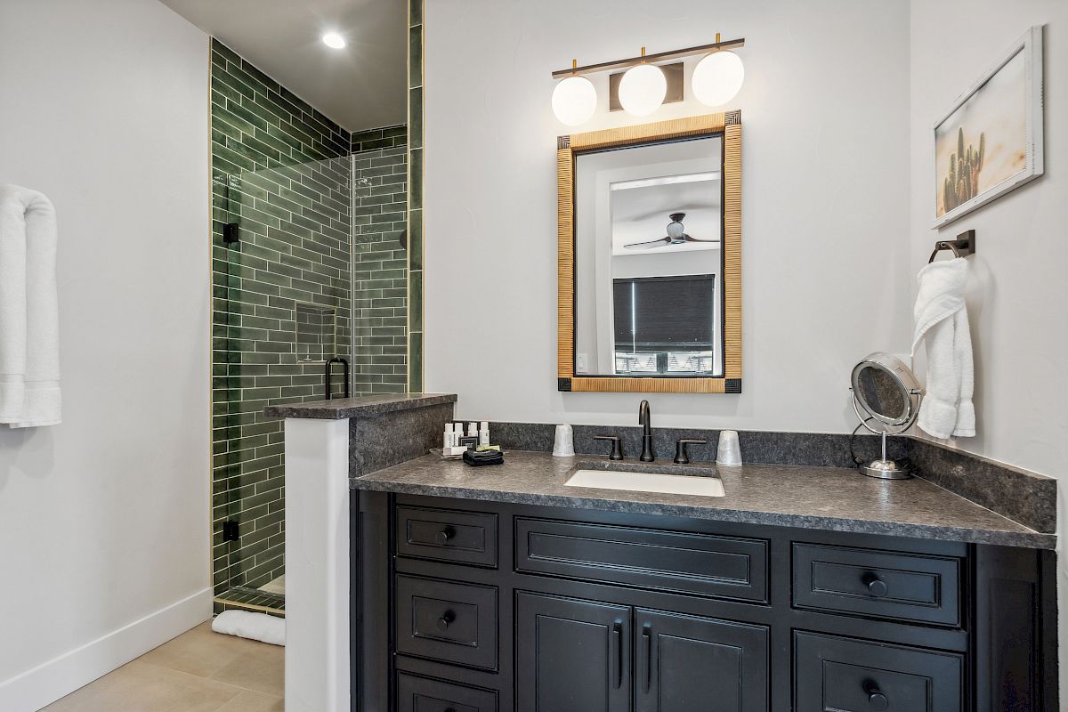 A modern bathroom with a dark vanity, mirror with lights, and a green-tiled shower area. White towels hang nearby.