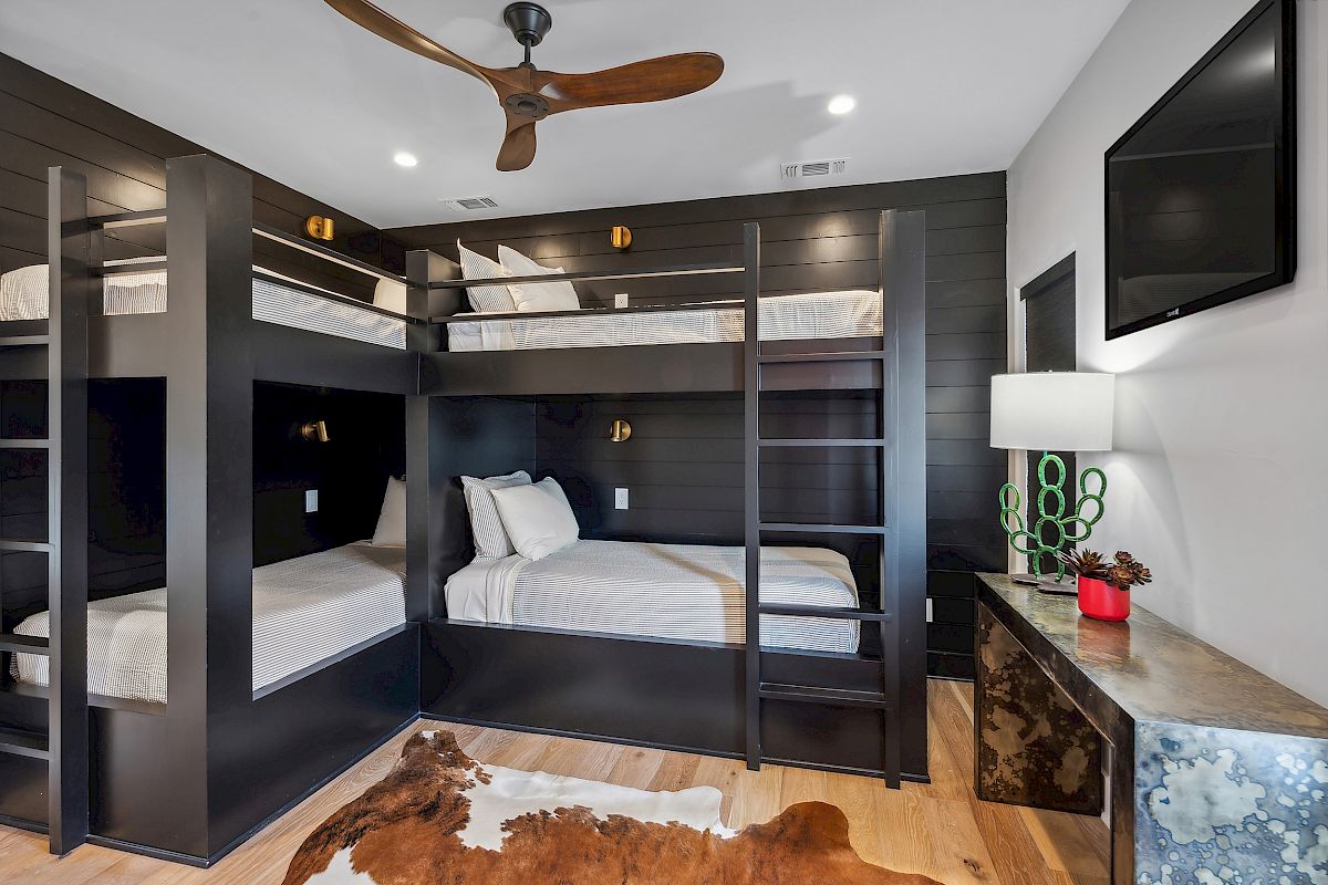 The image shows a room with black bunk beds, a ceiling fan, TV, and a cowhide rug. A dresser with a cactus lamp is also present.