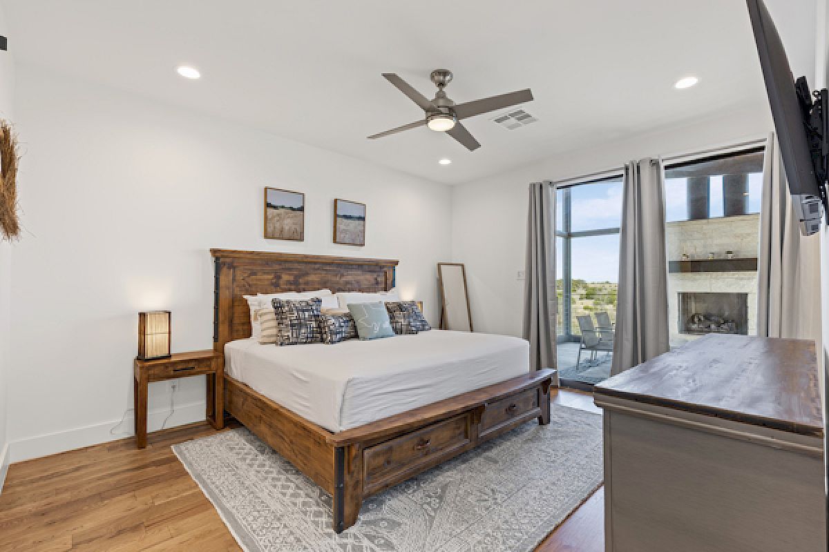A modern bedroom with a wooden bed, stylish decor, a ceiling fan, and large windows leading to an outdoor view complete the scene.