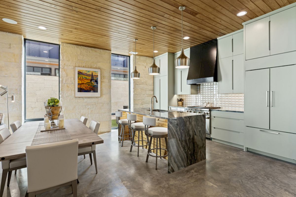 A modern kitchen features a marble island, dining table, pendant lights, and sleek cabinetry, with art on the wall and large windows.