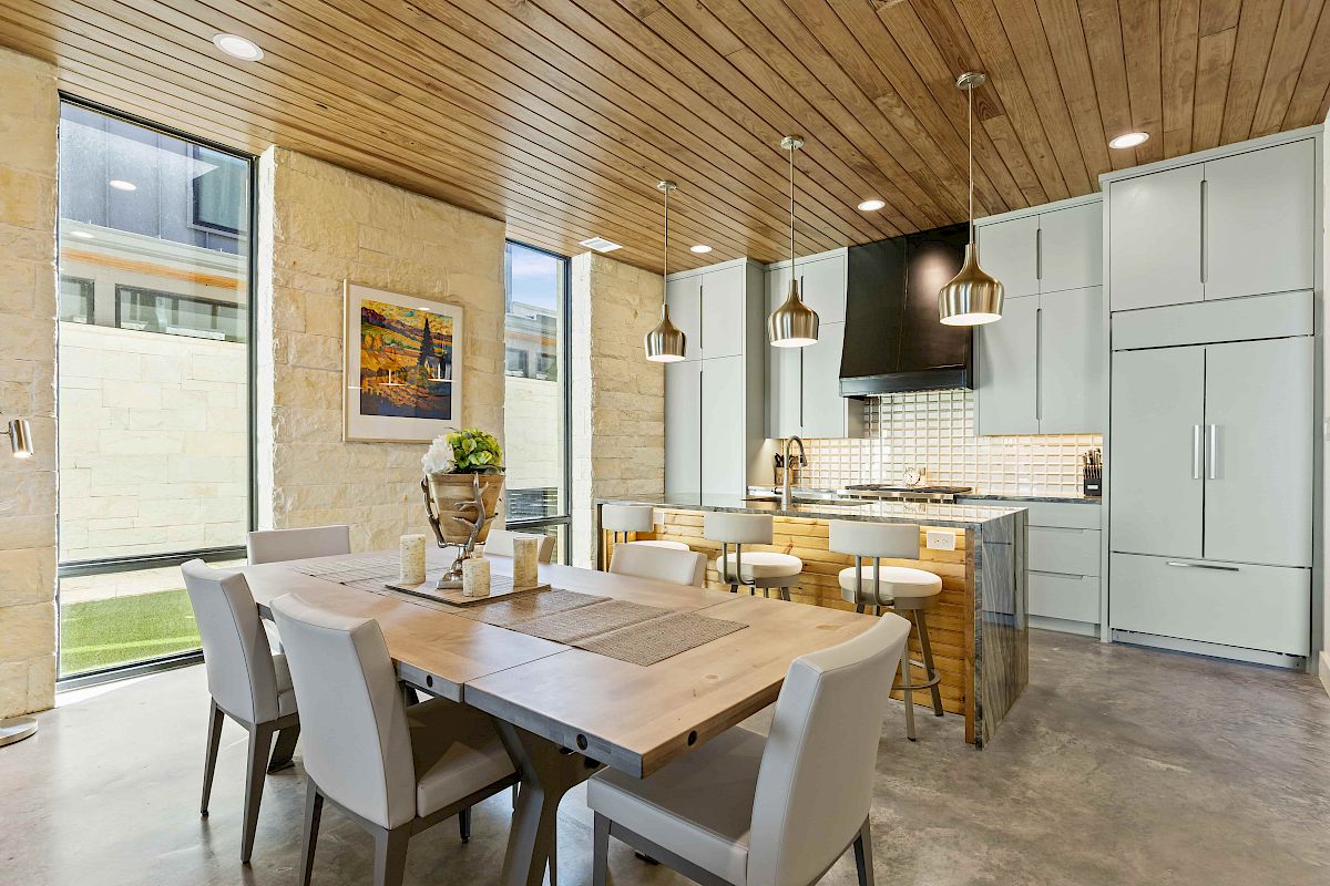 A modern kitchen and dining area with wood ceiling, pendant lights, large windows, and a wooden table with chairs.