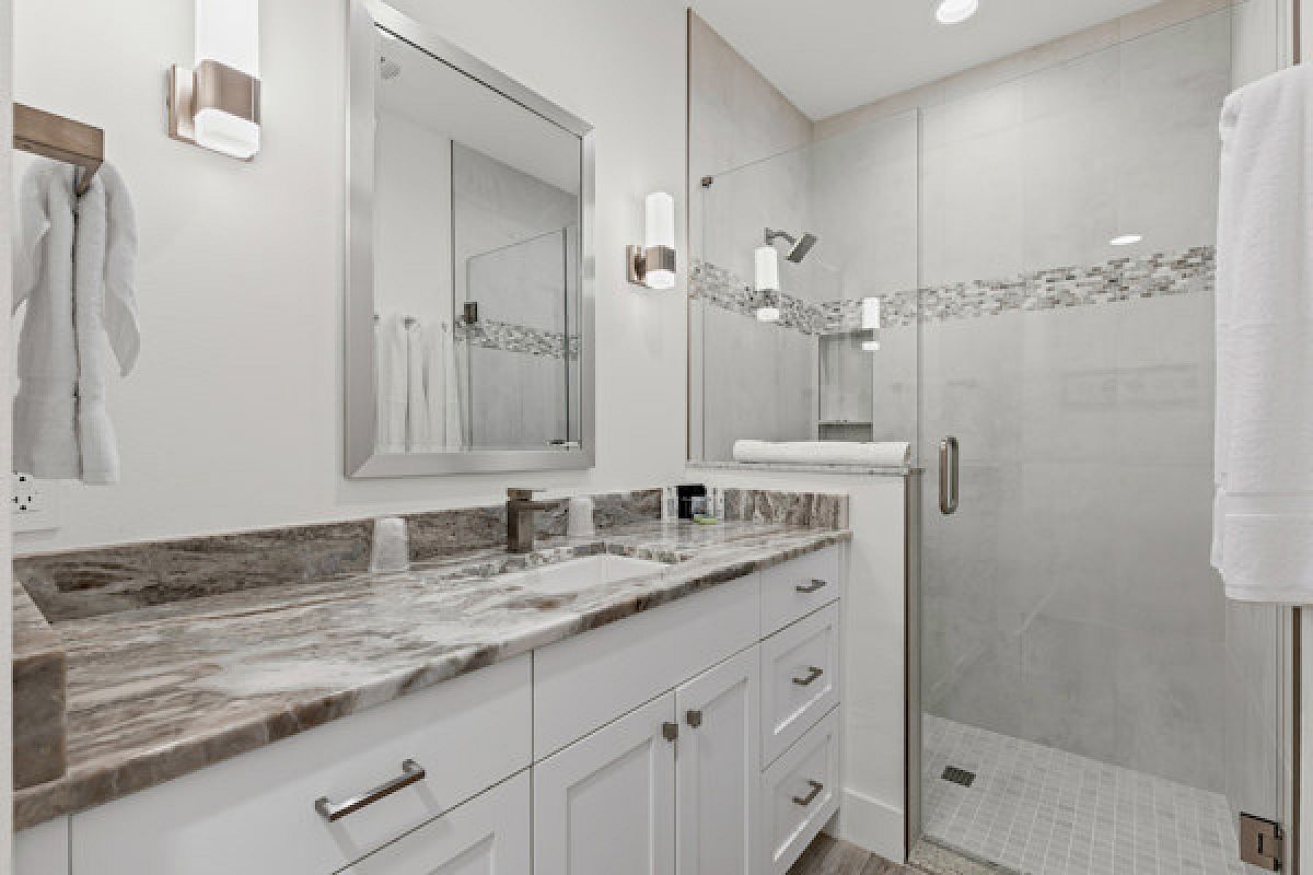 A modern bathroom with a marble countertop, glass-enclosed shower, white cabinets, wall lights, and a mirror above the sink.