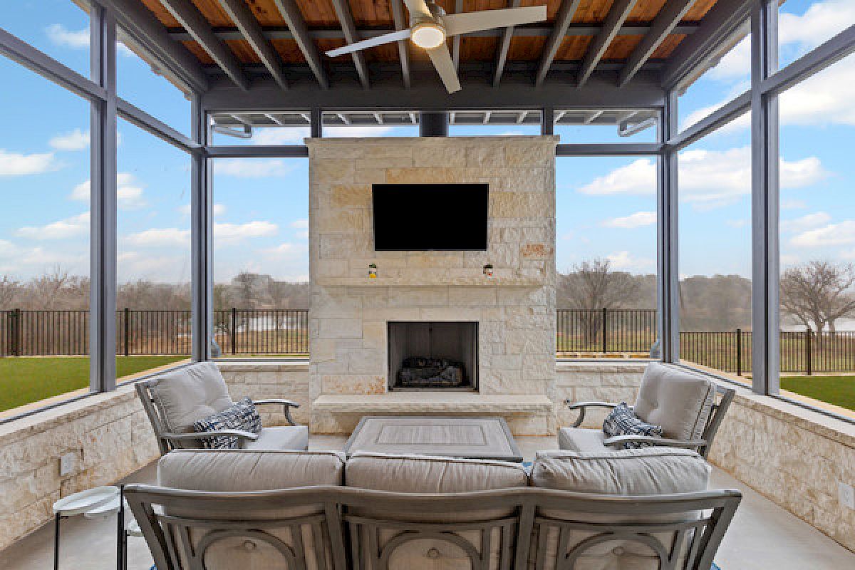 An outdoor seating area with a stone fireplace, TV, and ceiling fan, offering views of a fenced yard and a cloudy sky.