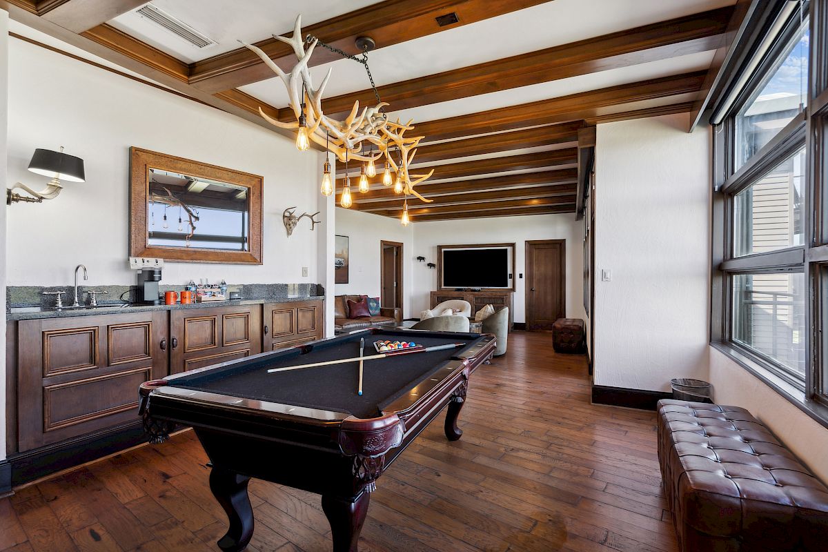 A stylish room features a pool table, antler chandelier, wooden beams, and a well-equipped bar area with a warm, rustic atmosphere.