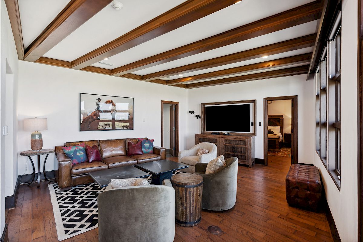 A cozy living room with wooden beams, leather sofa, armchairs, a large TV, and a painting on the wall, creating a warm atmosphere.