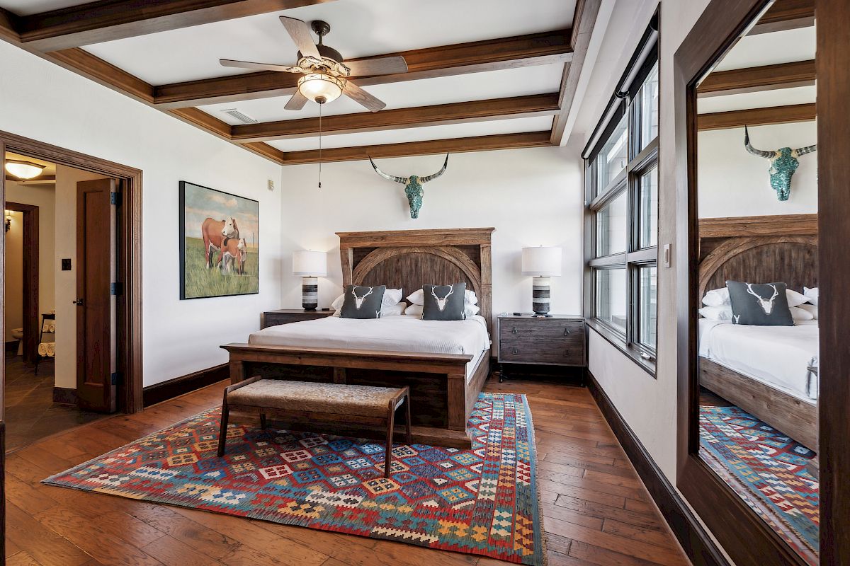 A cozy bedroom with a wooden bed, patterned rug, ceiling fan, and decorative art. Large windows and a mirror enhance the space.