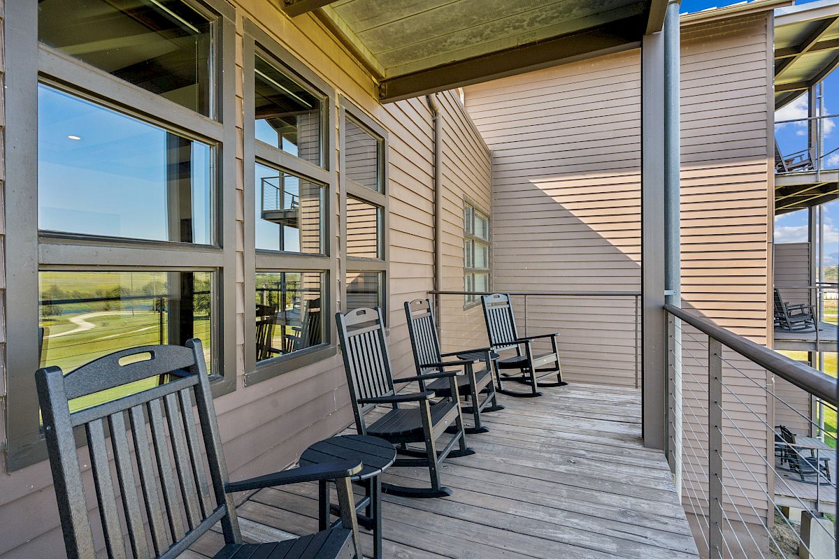 A balcony with wooden decking features several black rocking chairs and small round tables, overlooking an outdoor view through a railing.