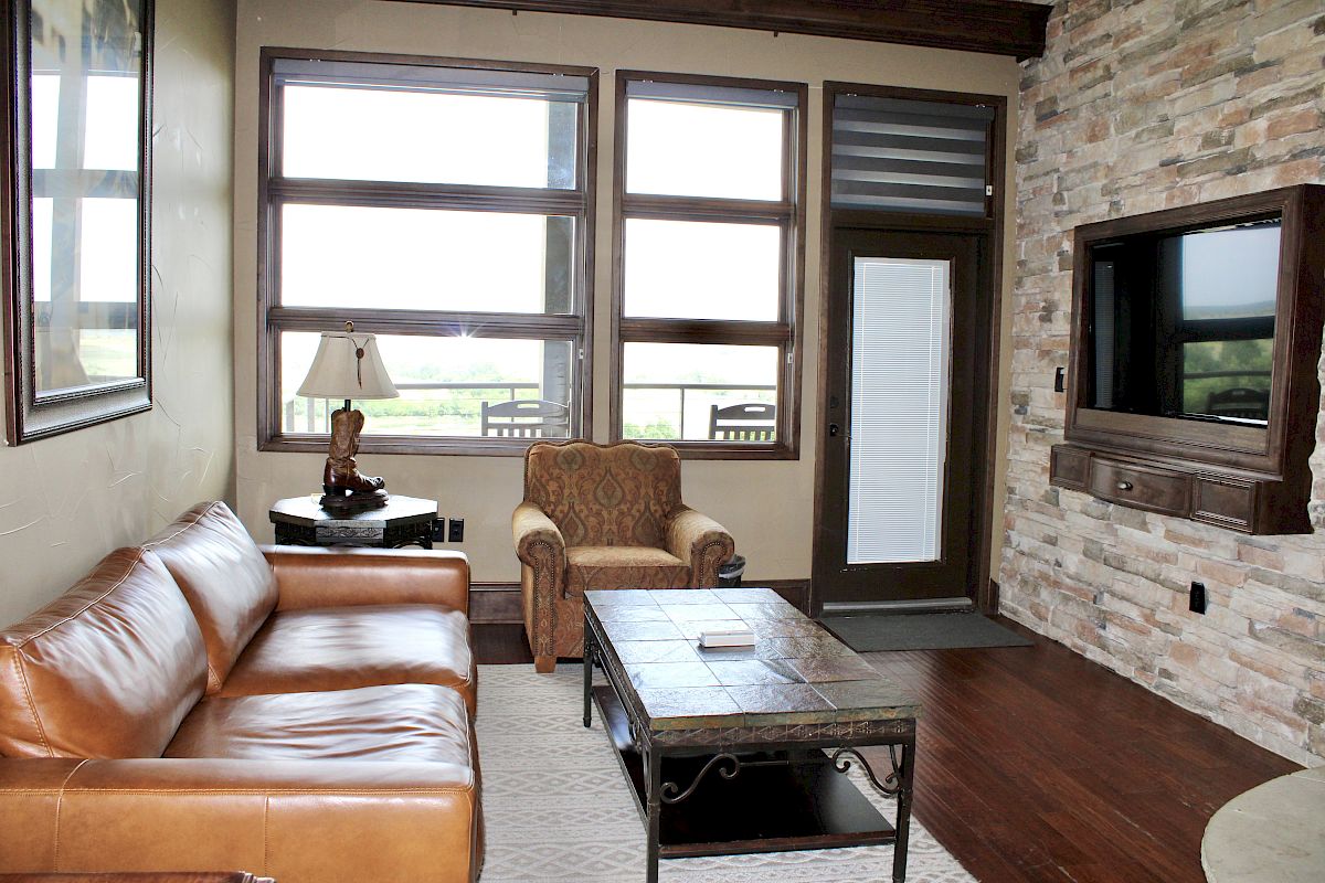 A cozy living room with a leather couch, armchair, coffee table, stone wall, TV, and large windows with a scenic view.