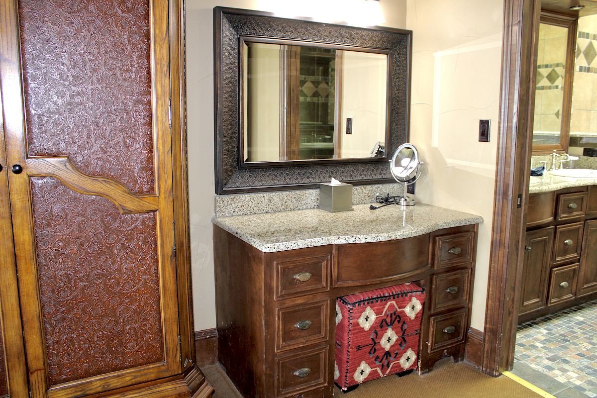 The image shows a bathroom vanity with a mirror, wooden cabinets, a granite countertop, and a decorative ottoman underneath.