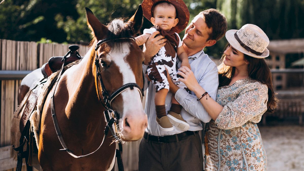 A family with a baby wearing a cowboy hat is standing next to a horse in a sunny outdoor setting.
