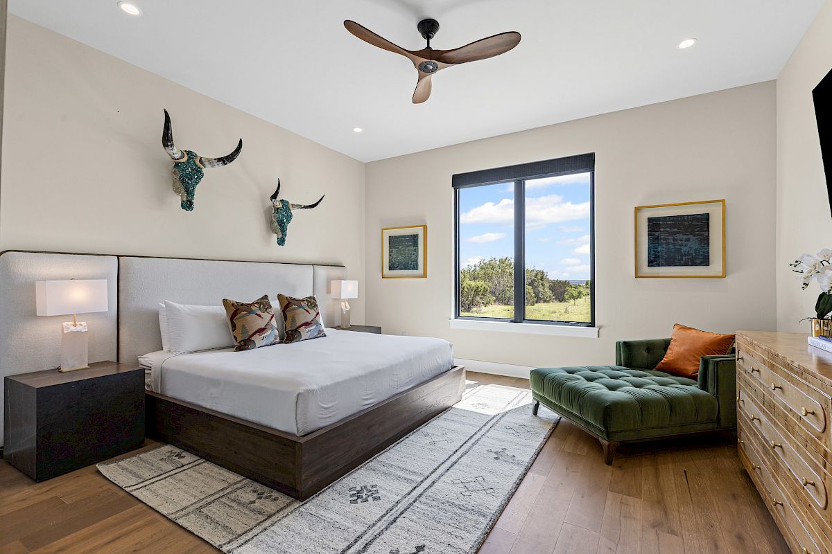 A stylish bedroom with a bed, green chaise lounge, wooden dresser, window view, decorative wall art, and ceiling fan.