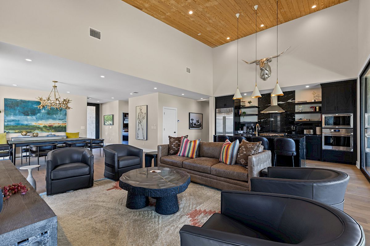 The image shows a modern living room with a high ceiling, stylish furniture, a kitchen area, and a dining section with artwork.