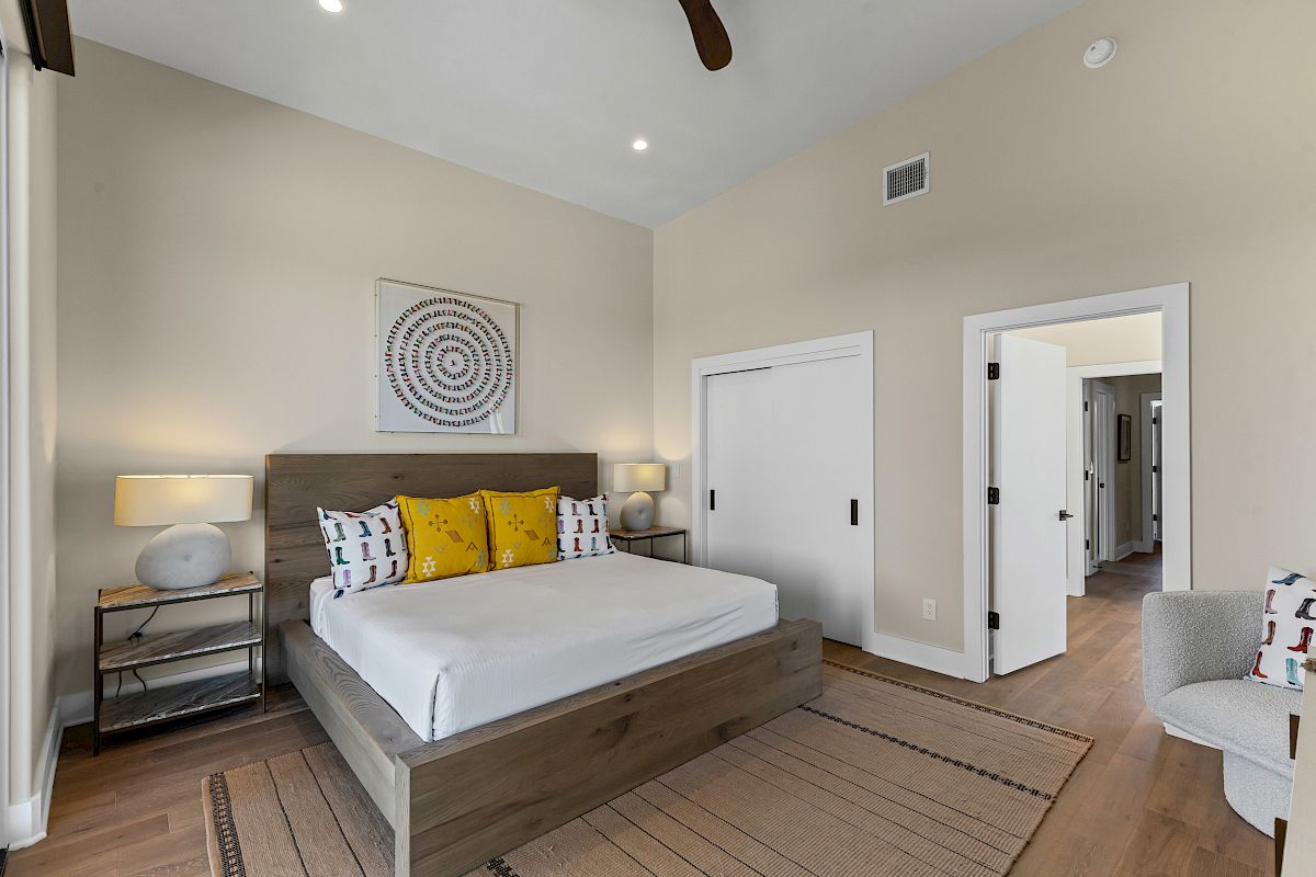 A modern bedroom with a wooden bed, patterned pillows, nightstands, lamps, and abstract wall art, with beige walls and wooden flooring.