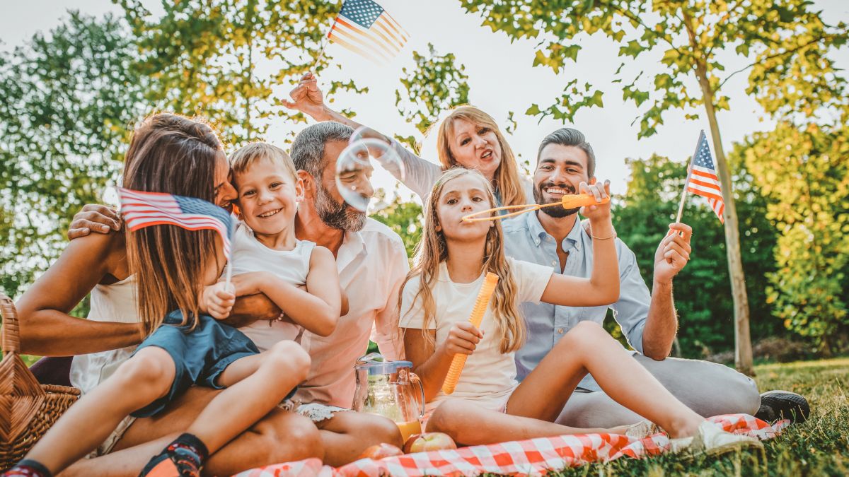 A group of people having a picnic on a sunny day, waving flags and blowing bubbles, enjoying time together on a checkered blanket outdoors.