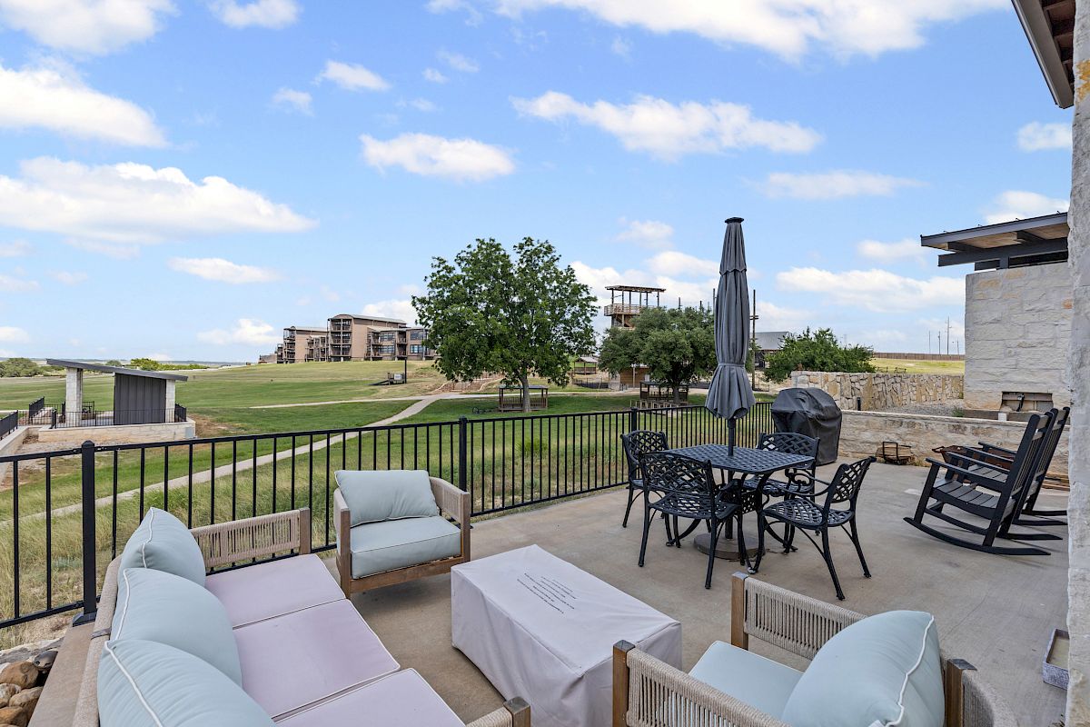 A patio with outdoor seating, a table, an umbrella, and a grill overlooks a grassy area and buildings under a partly cloudy sky.