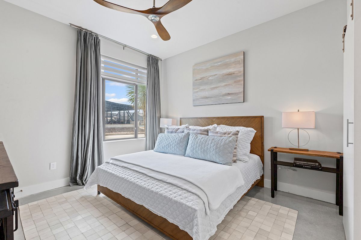 A modern bedroom with a bed, artwork, window with gray curtains, ceiling fan, bedside table, lamp, and a soft rug.