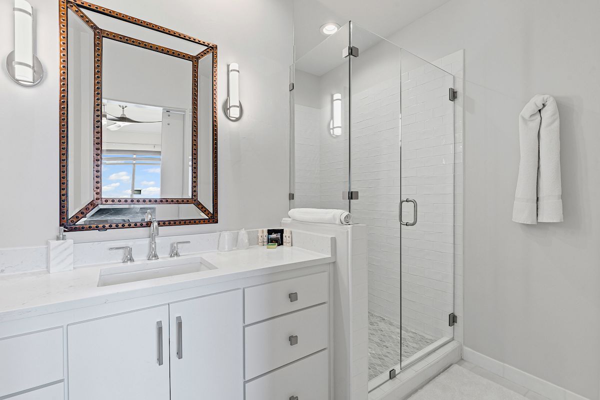 The image shows a modern white bathroom with a large mirror, a double sink vanity, and a glass-enclosed shower area.