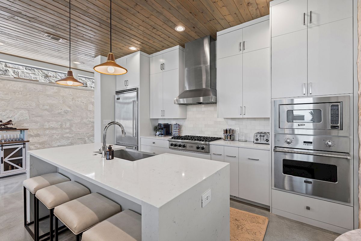 Modern kitchen with white cabinets, stainless steel appliances, island with sink, stools, pendant lights, and wooden ceiling.