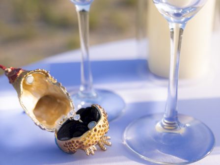 An ornate, gold shell-shaped box with rings inside is next to two clear wine glasses on a table.