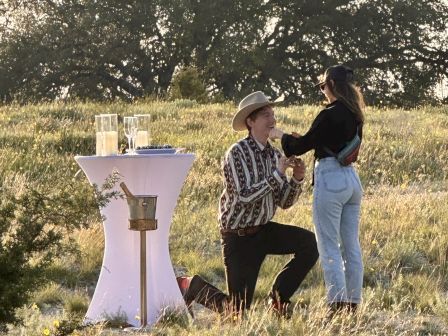 A person kneels, holding a ring to another person in a grassy field. A small table with a decorative cloth and items is nearby.