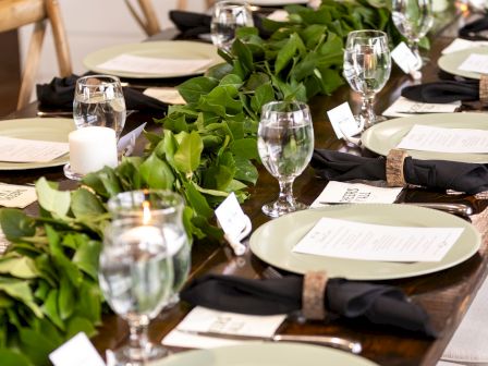 A long, elegantly set dining table with plates, black napkins, glassware, menus, and a green leaf centerpiece decorates the scene.