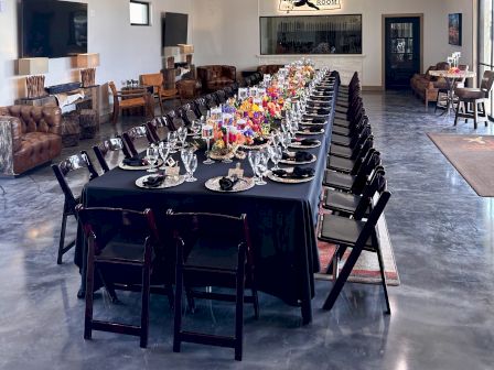 A long dining table is set with chairs, cutlery, and flower arrangements, under hanging greenery and disco balls in a modern room.