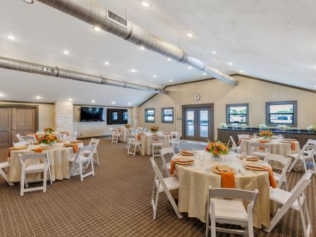 The image shows a decorated banquet room with round tables, white chairs, and orange accents, ready for an event or gathering.