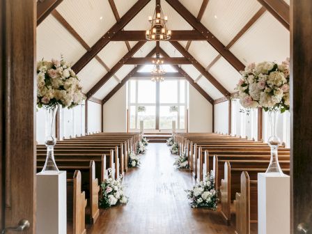 A beautifully decorated chapel interior with wooden pews, floral arrangements, and large windows at the front allowing natural light to flood in.