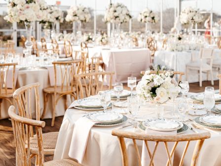 The image shows an elegant table setting in a tent with white flowers and wooden chairs, designed for a formal event or wedding.